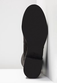 Semelle de chaussure noire avec motifs de grip texturés et une petite marque de taille, inclinée contre une surface blanche d'angle.