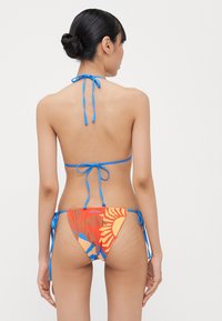 Bikini s halter vratnim oblikom, živo oranžne tkanine z vzorci sonca in palm, modrimi vezmi in minimalno pokritostjo hrbta.