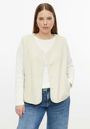 Femme aux cheveux châtains raides porte une veste polaire crème à boutons par-dessus une chemise blanche à manches longues et un jean bleu, debout devant un fond blanc.