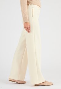 Pantaloni a gamba larga in tessuto crema, caratterizzati da una texture liscia, pince e vita alta. Indossati con mocassini di colore chiaro.