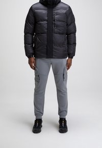 Doudoune noire avec une finition lisse et un col montant, associée à un pantalon de jogging gris avec poches zippées et jambes fuselées. Des baskets noires complètent le look.