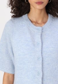 Cardigan à manches courtes bleu clair en tissu doux, avec un col rond et cinq boutons assortis sur le devant.