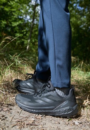 Schwarze Sportschuhe mit strukturiertem Mesh-Obermaterial, Schnürsenkeln und vier grauen Streifen; kombiniert mit blauen Sporthosen und grauen Socken.