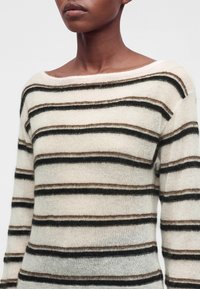 Personne portant un pull en tricot blanc avec des rayures horizontales noires et marron et un large col bateau sur un fond uni.