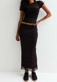 Schwarzes, transparentes Oberteil mit kurzen Ärmeln, kombiniert mit einem schwarzen Floraltrock, der rote Kirschmuster und einen fransigen Saum hat. Schwarze Sneaker sind enthalten.