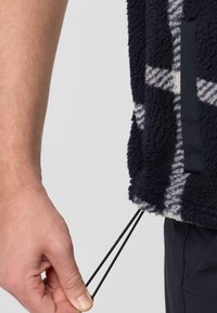 Flauschige navyblaue Fleecejacke mit einem weißen karierter Muster. Verfügt über verstellbare Kordeln am Saum. Hand, die die Schnur greift.