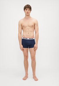 Ναυτικές μποξερί briefs με ζώνη λογότυπου Moschino. Διαθέτουν εφαρμογή που αγκαλιάζει το σώμα και λεία υφή, σχεδιασμένα για άνεση. Μοντέλο που στέκεται ξυπόλυτο, χωρίς μπλούζα.