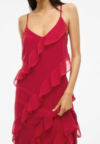 Raudona šifono suknelė su V formos iškirpte, plonais petnešomis ir sluoksniuotomis volanomis priekyje. Lygi tekstūra ir laisvas siluetas.