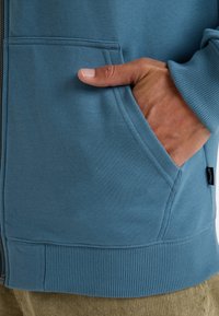 Blue sweatshirt with ribbed cuffs and hem, featuring a zip closure and side pockets. Soft texture and casual design visible.