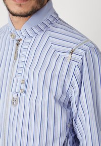 Camisa de rayas en azul claro y blanco. Presenta un cuello alto, cierre frontal y bolsillo con cremallera lateral. Tejido ligero con textura suave.