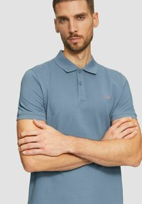 Mann mit kurzen Haaren und Bart, der ein blaues kurzärmliges Polohemd trägt und mit verschränkten Armen vor einem einfarbigen Hintergrund steht.