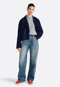 Giacca navy cropped su un maglione grigio, abbinata a jeans in denim blu a gamba larga e scarpe rosse. Gioielli semplici completano il look.