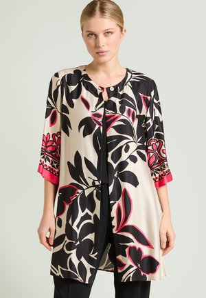 Femme portant un long chemisier beige avec de grandes fleurs noires et roses, manches trois-quarts et encolure ronde avec détail de boutons.