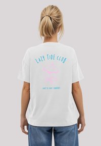 Weißes Baumwoll-T-Shirt mit einem bunten Grafikdesign auf der Rückseite. Der Text lautet "Lazy Tide Club" in Blau, begleitet von einem pinken Strandschirm und einem Stuhl.