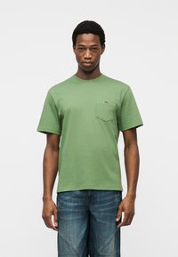 Νεαρός άνδρας που φοράει ανοιχτό πράσινο t-shirt με τσέπη και μπλε τζιν, στέκεται μπροστά σε απλό φωτεινό φόντο.