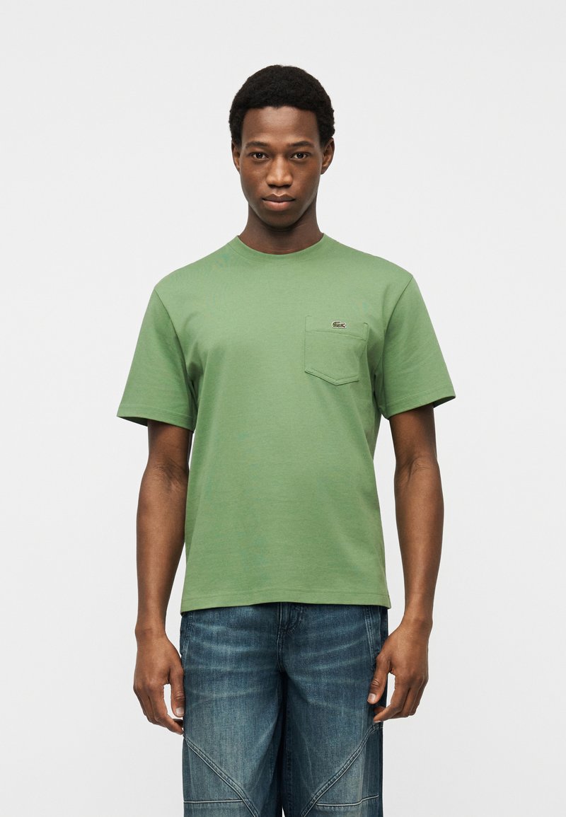 Νεαρός άνδρας που φοράει ανοιχτό πράσινο t-shirt με τσέπη και μπλε τζιν, στέκεται μπροστά σε απλό φωτεινό φόντο.