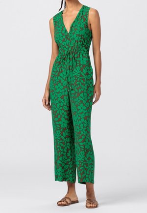 Kobieta ubrana w bezrękawnikowy zielony kombinezon z dużym ciemnym kwiatowym wzorem i ściągaczem w talii, zestawiony z brązowymi, płaskimi sandałami.