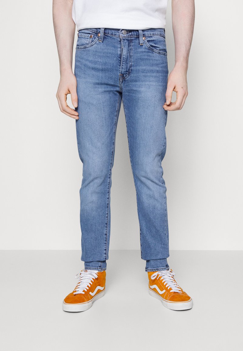 Világoskék mosású farmernadrág, slim fit szabásban, öt zsebbel, finom kopásnyomokkal, klasszikus gombos és cipzáras záródással. Narancssárga sportcipővel párosítva.