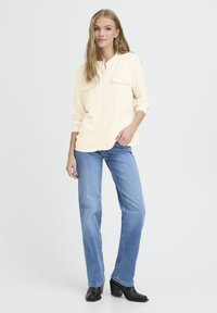 Light beige button-up shirt with two chest pockets, paired with blue straight-leg jeans and black heeled boots. Smooth texture, casual fit.