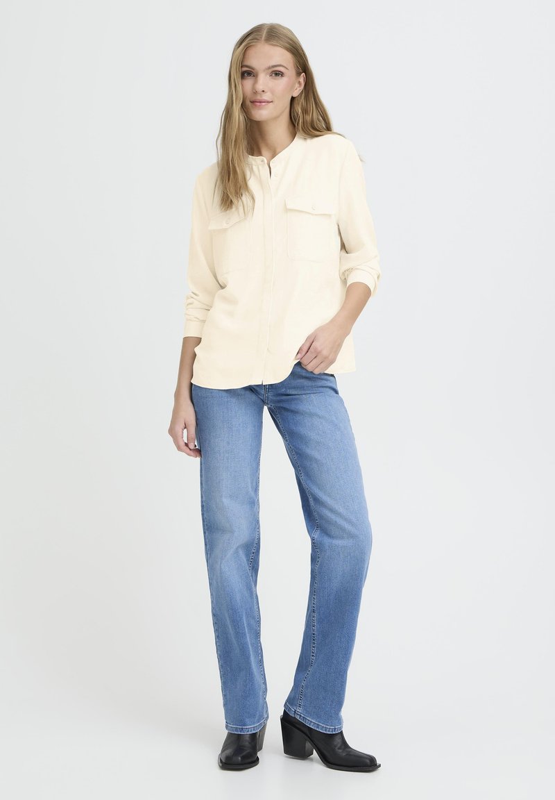 Light beige button-up shirt with two chest pockets, paired with blue straight-leg jeans and black heeled boots. Smooth texture, casual fit.