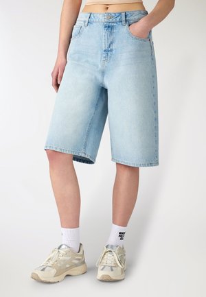 Persona che indossa pantaloncini di jeans alla moda sotto il ginocchio di colore azzurro chiaro, calzini bianchi alla caviglia e scarpe da ginnastica beige e argento, in piedi su uno sfondo semplice.