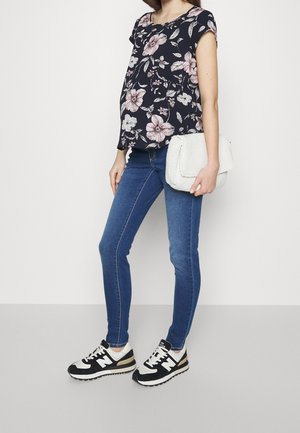 Kobieta w czarnej kwiecistej bluzce, niebieskich obcisłych dżinsach, czarno-białych trampkach, trzymająca przy boku teksturowaną białą torebkę kopertówkę.