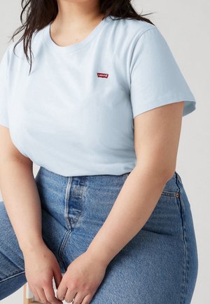 Personne portant un t-shirt Levi's bleu clair et un jean bleu taille haute, assise avec les mains reposant sur les genoux, devant un fond neutre.