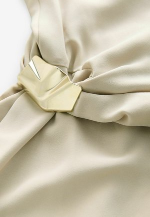 Beżowa tkanina o gładkiej teksturze, zebrana w centrum, z złotym metalicznym zapięciem o geometrycznym wzorze.