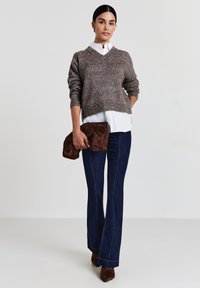 Maglione marrone lavorato a maglia con scollo a V, camicia bianca sotto, jeans svasati blu scuro e borsa a tracolla in suede marrone. Stivali col tacco marroni.
