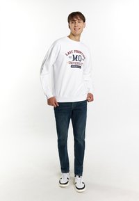 Hvid sweatshirt med rød og navy tekst "LAST FRONTIER MO UNIVERSITY", parret med mørkeblå jeans og sort-hvide sko.