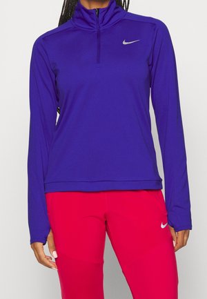 Fioletowa sportowa bluzka z długim rękawem, zamkiem na ćwiartkę i wysokim kołnierzem. Na lewej piersi znajduje się małe srebrne logo Nike.