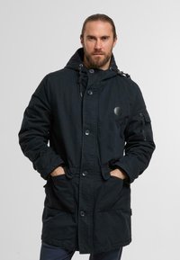 Black parka jacket with a hood, large front pockets, button closure, and adjustable drawstrings. Features a logo patch on the chest.