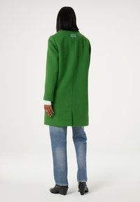 Groene wollen jas met een op maat gemaakt ontwerp, halflang, met een enkele achteropening en geknipte revers. Draag het over lichtblauwe jeans en zwarte hakken.