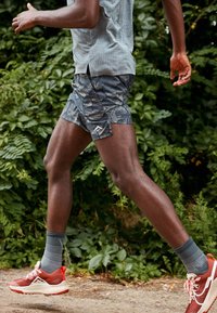 Person som springer utomhus på en grusstig och bär mönstrade shorts, grå strumpor och röda löparskor med grönska i bakgrunden.