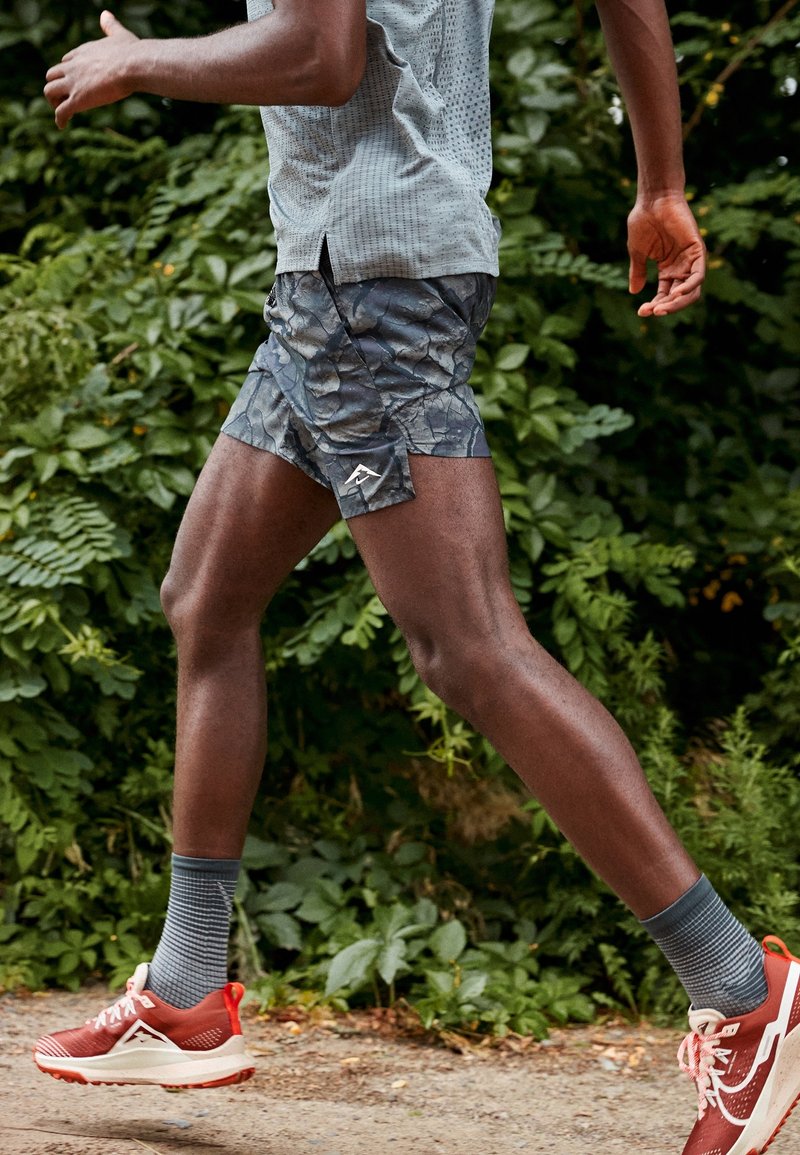 Person som springer utomhus på en grusstig och bär mönstrade shorts, grå strumpor och röda löparskor med grönska i bakgrunden.