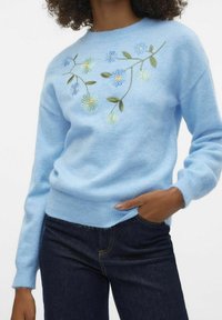 Light blue knit sweater features floral embroidery in shades of blue and green on the front, with ribbed cuffs and hem. Worn with dark jeans.