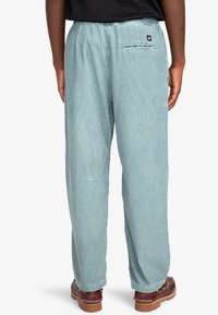 Light blue, loose-fitting pants with an elastic waistband. Features a back pocket with a zipper and textured fabric. Worn with brown shoes.
