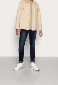 Beige bomullsjacka med framfickor, en ljusbeige t-shirt, mörka slim-fit jeans och vita sneakers med grå detaljer och ljusbruna sulor.