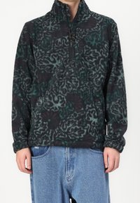 Osoba ubrana w czarny i zielony polar w wzory z ćwierć zamkiem i jasnoniebieskie luźne dżinsy.