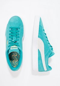 Turkusowe zamszowe sneakersy z białymi akcentami, płaskimi sznurowadłami i teksturowaną gumową podeszwą; mają wzorzyste wnętrze. Logo widoczne na języku.