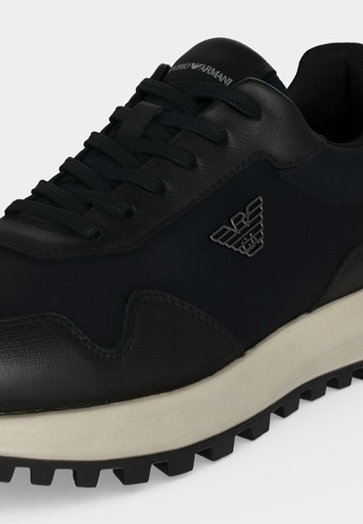 Baskets basses noires en cuir texturé, lacets noirs, semelle blanche et logo métallique de l'aigle Armani sur le côté.