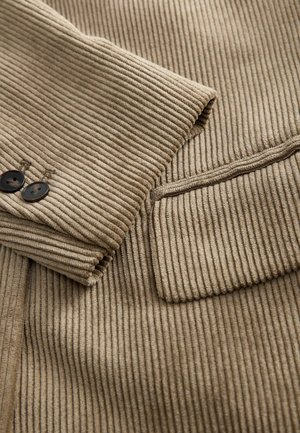Beige kordfløjljakkeærme med to mørke knapper og detaljer med klaplomme på ribbet tekstiler.