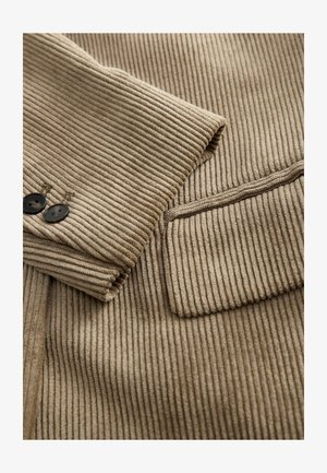 Ärmel einer beigen Cordjacke mit zwei dunklen Knöpfen und einem Klappentaschen-Detail auf dem gerippten Textil.