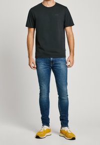Uomo che indossa una maglietta nera a maniche corte, jeans slim-fit blu e sneaker giallo-bianche, in piedi di fronte a uno sfondo neutro.
