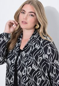 Černá bunda s bílým květinovým vzorem, otevřený přední díl, dlouhé rukávy. Zlaticové náušnice s dramatickým designem dodávají kontrast. Hladká textura látky.