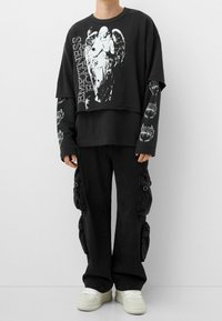 Černá oversized mikina s grafickým potiskem, nošená přes dlouhý rukáv. Černé cargo kalhoty s nařasenými detaily a bílé tenisky.
