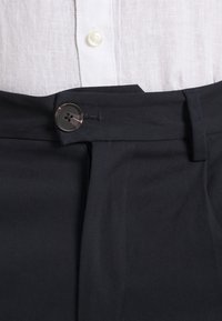 Bílá košile s knoflíky zasunutá do černých kalhot s vysokým pasem, které jsou zapnuté jediným tmavým knoflíkem a mají viditelný poutko na opasek.