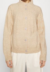 Cardigan en tricot beige avec un col montant, featuring cinq boutons ornés et des bords côtelés aux poignets et à l'ourlet. Texture douce, coupe décontractée.