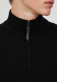 El suéter negro de cremallera presenta un cuello alto con textura acanalada y un patrón en forma de chevrón, acentuado por un pequeño logo en el tirador de la cremallera.