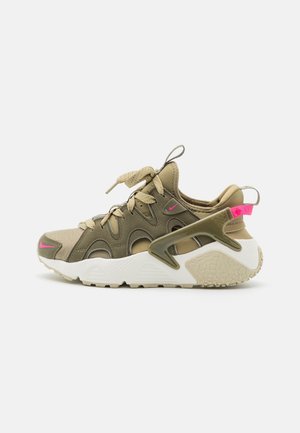 Beige og olivengrøn atletiksko med hvid sål, pink Nike-logoer, snørefront og hælstøttebur på en neutral baggrund.